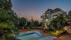 A swimming pool with a red bench, surrounded by lush greenery and a multi-story building with balconies. - CosmicStays Venus Villa- Private pool  (Mawal)