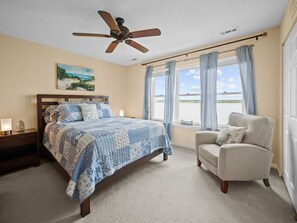 Quarto com cama, poltrona, ventilador de teto, quadro e janela com vista para a praia.