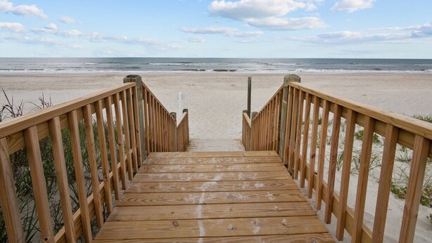 Ein Holzsteg führt zu einem Strand, im Hintergrund leuchtender Himmel und Ozean.