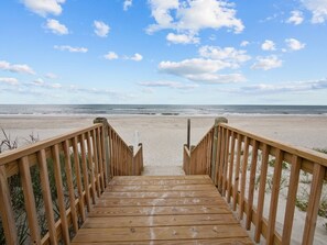 A wooden boardwalk leading to a beach with a clear sky and ocean in the background.