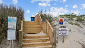 Eine Holztreppe führt zu einem Sandstrand, an dem mehrere Schilder stehen. Eines betrifft North Topsail Beach und verlangt, dass Hunde angeleint sind und Kot entfernt wird. Ein weiteres Schild warnt vor Beschädigungen der Dünen und kündigt eine Strafe bei Nichtbeachtung an.