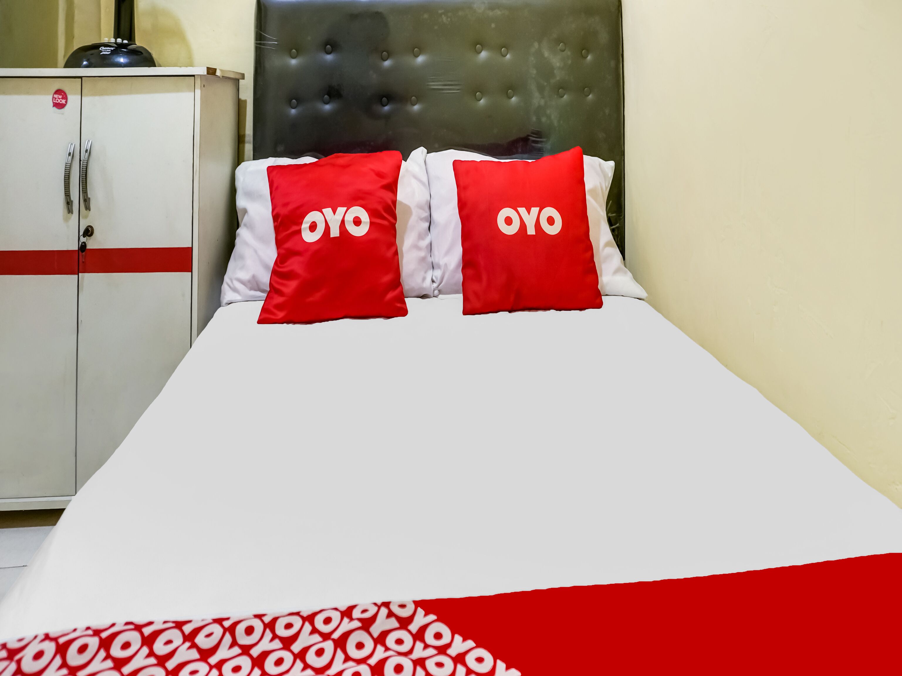 Une chambre d’hôtel avec un lit, deux oreillers rouges ornés du logo « OYO », une couverture blanche et rouge à motifs et une armoire blanche avec une bande rouge.