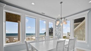 A dining area with a white table and chairs, large windows with a view of the ocean, and a ceiling light fixture. - Island Drive 4180 (North Topsail Beach)