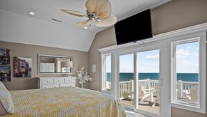 A bedroom with a bed, a ceiling fan, a TV, a dresser, a mirror, and a balcony with a view of the ocean. - Island Drive 4180 (North Topsail Beach)