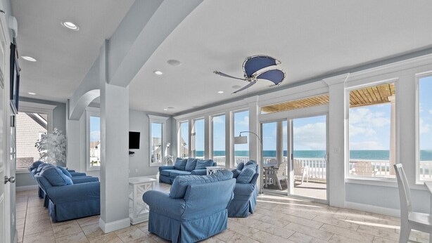 A spacious living room with a large window overlooking the ocean, a ceiling fan, and a seating area with blue sofas. - Island Drive 4180 (North Topsail Beach)