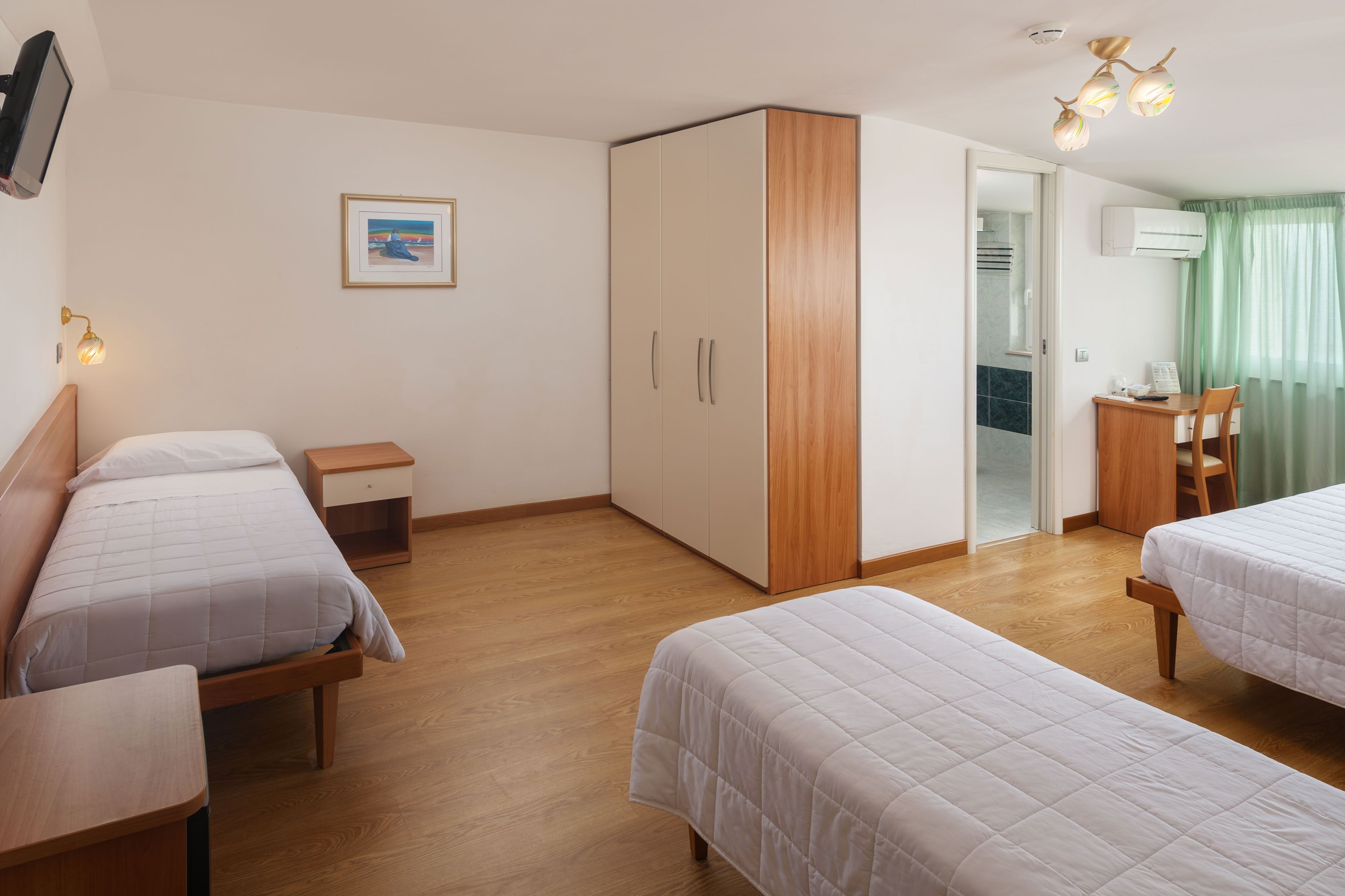 Une chambre d’hôtel avec deux lits simples, une armoire en bois, un bureau avec une chaise, une petite table de chevet et un téléviseur fixé au mur.