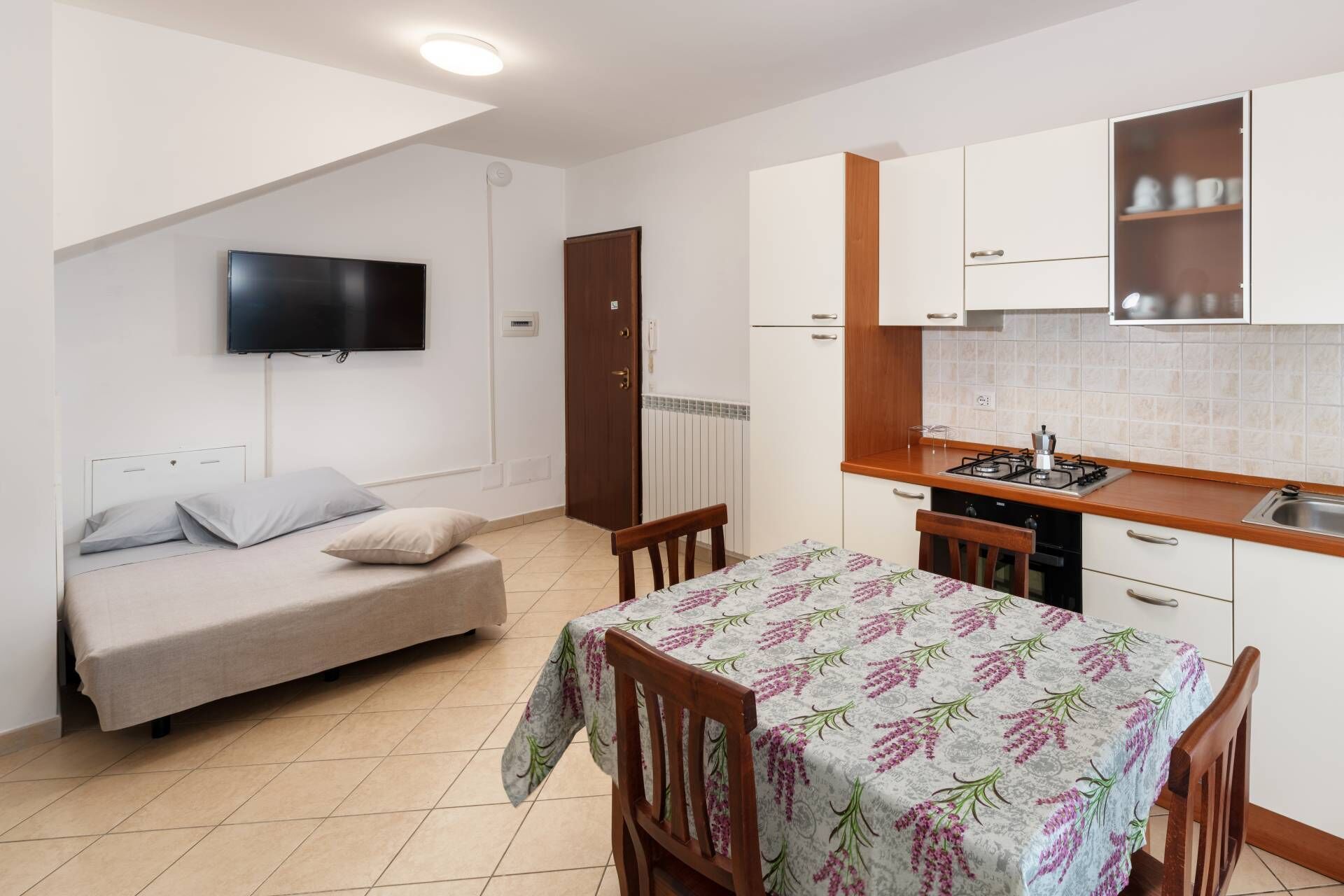 A compact room with a bed, a dining table, a kitchen area, and a TV mounted on the wall.
