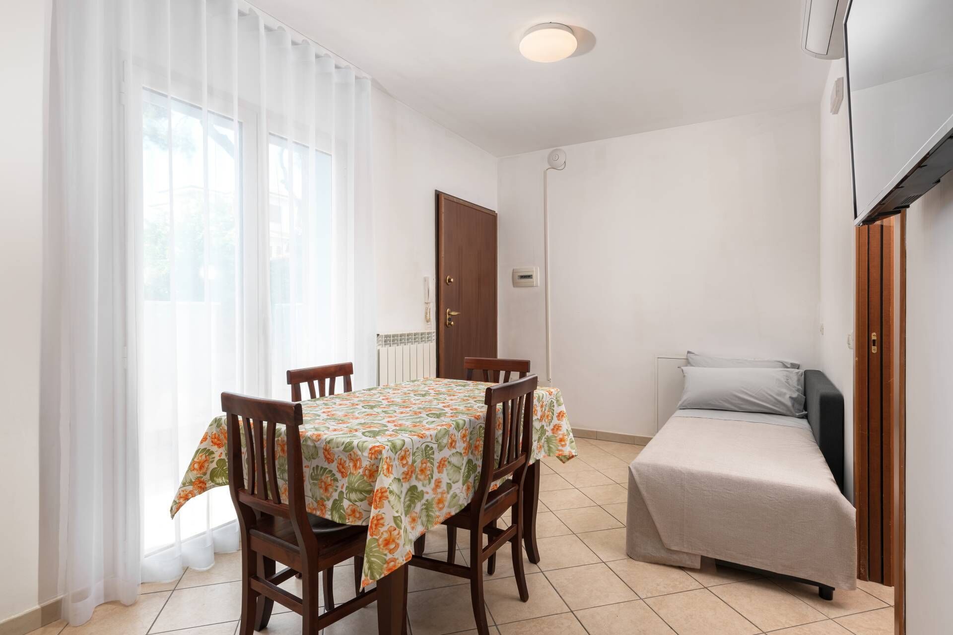 Una habitación pequeña y bien iluminada, con una mesa de comedor y sillas, una cama con colchón gris, y un radiador en la pared.