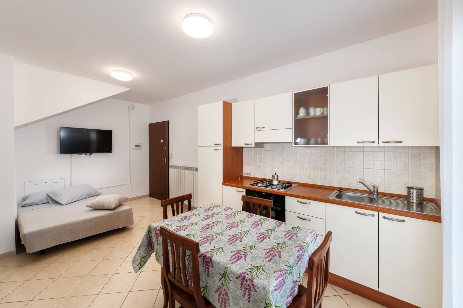 A compact kitchen with white cabinets, a dining table with a floral tablecloth, a built-in stove, and a TV mounted on the wall.
