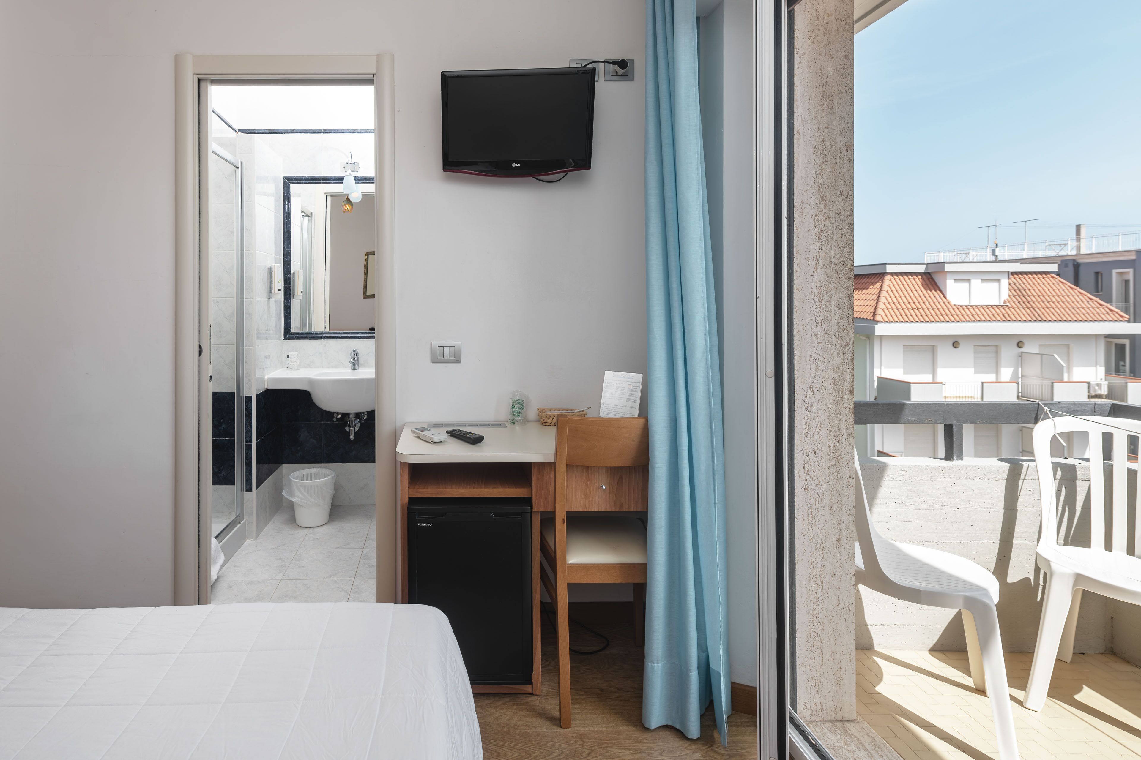 Habitación de hotel pequeña con cama, escritorio con silla, televisión montada en la pared y balcón con vistas a edificios.