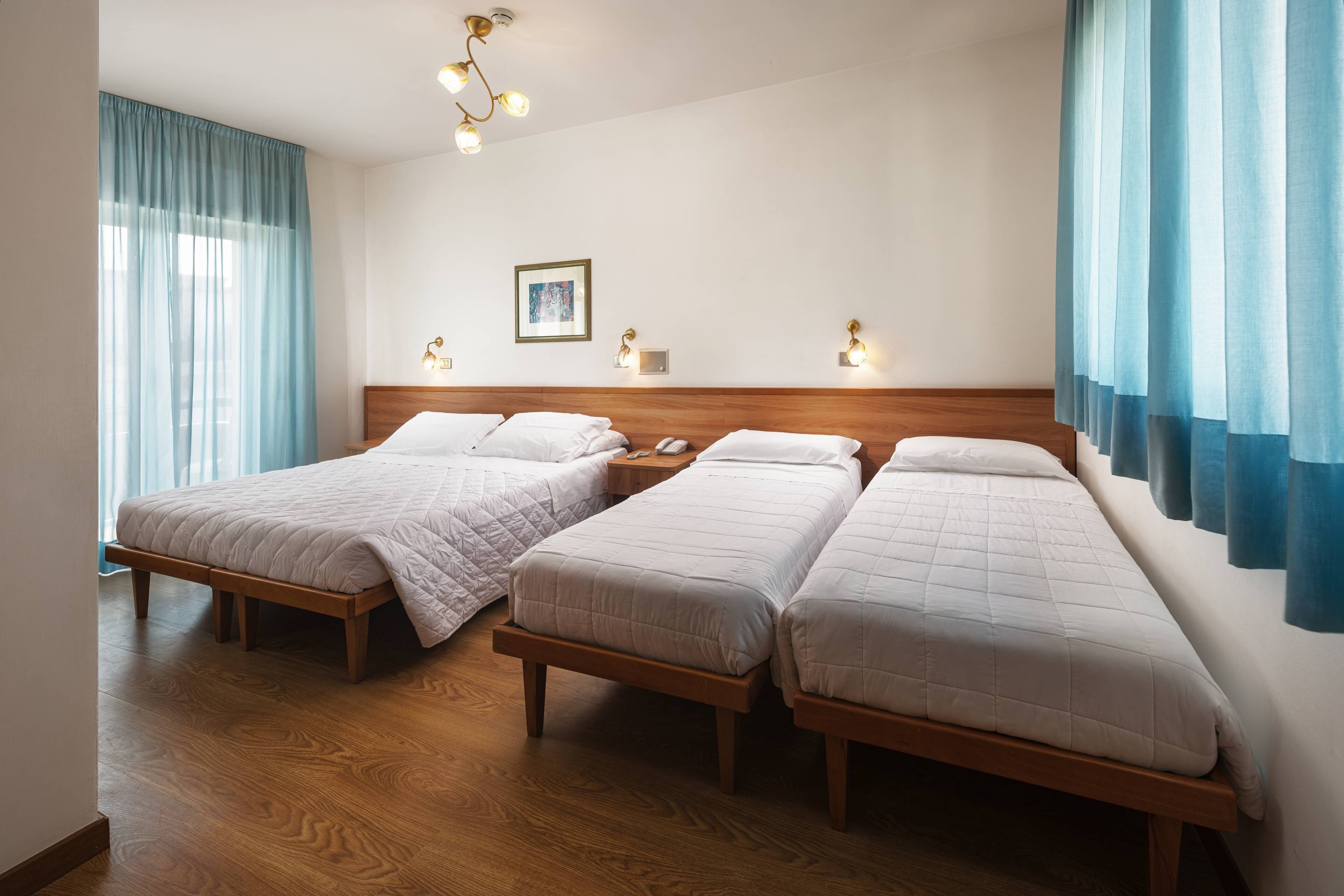 Habitación de hotel con tres camas individuales, cabeceros de madera, suelo de madera, una lámpara fijada a la pared y un cuadro enmarcado.