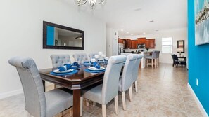 A modern dining area with a wooden table and grey upholstered chairs, a kitchen in the background, and a blue accent wall.