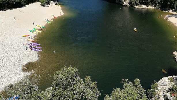 A natural rock arch over a river with a sandy beach and kayaks lined up on the shore. - MONLOUBIER (Villeneuve-de-Berg)