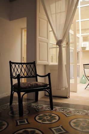 A chair with a patterned cushion in a room with patterned floor tiles, a white door with a curtain, and a view of an adjacent room.