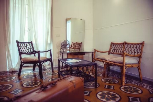 A room with two wicker chairs, a small table, a mirror, and a suitcase on a patterned floor.