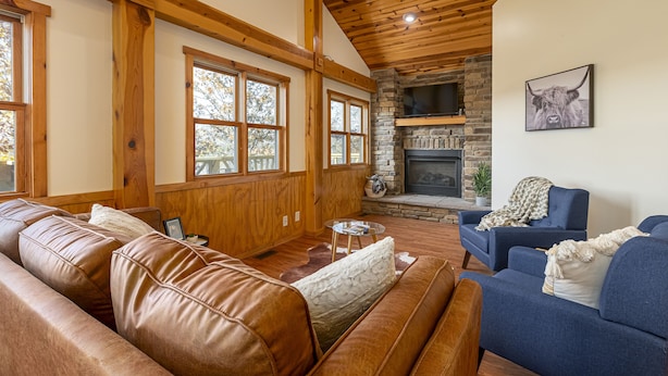 A cozy living room with leather sofas, a stone fireplace, and large windows.