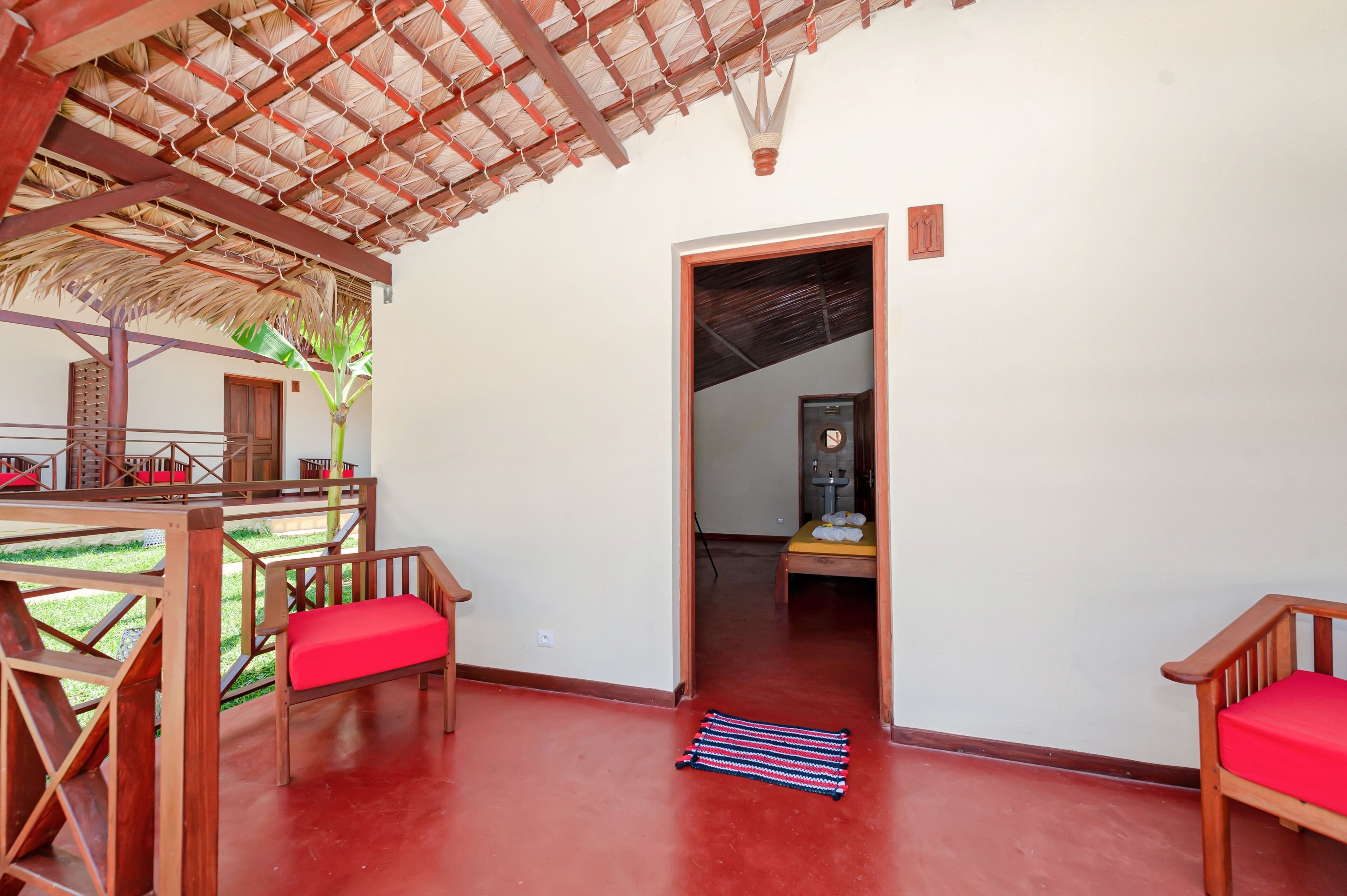 A room with a red floor, wooden furniture, a thatched roof, and a door leading to another room.