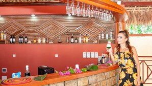 A bar area with a wooden wall, bottles of alcohol, and a woman standing by the counter. - BLUE SKY RESORT (Nosy Be)