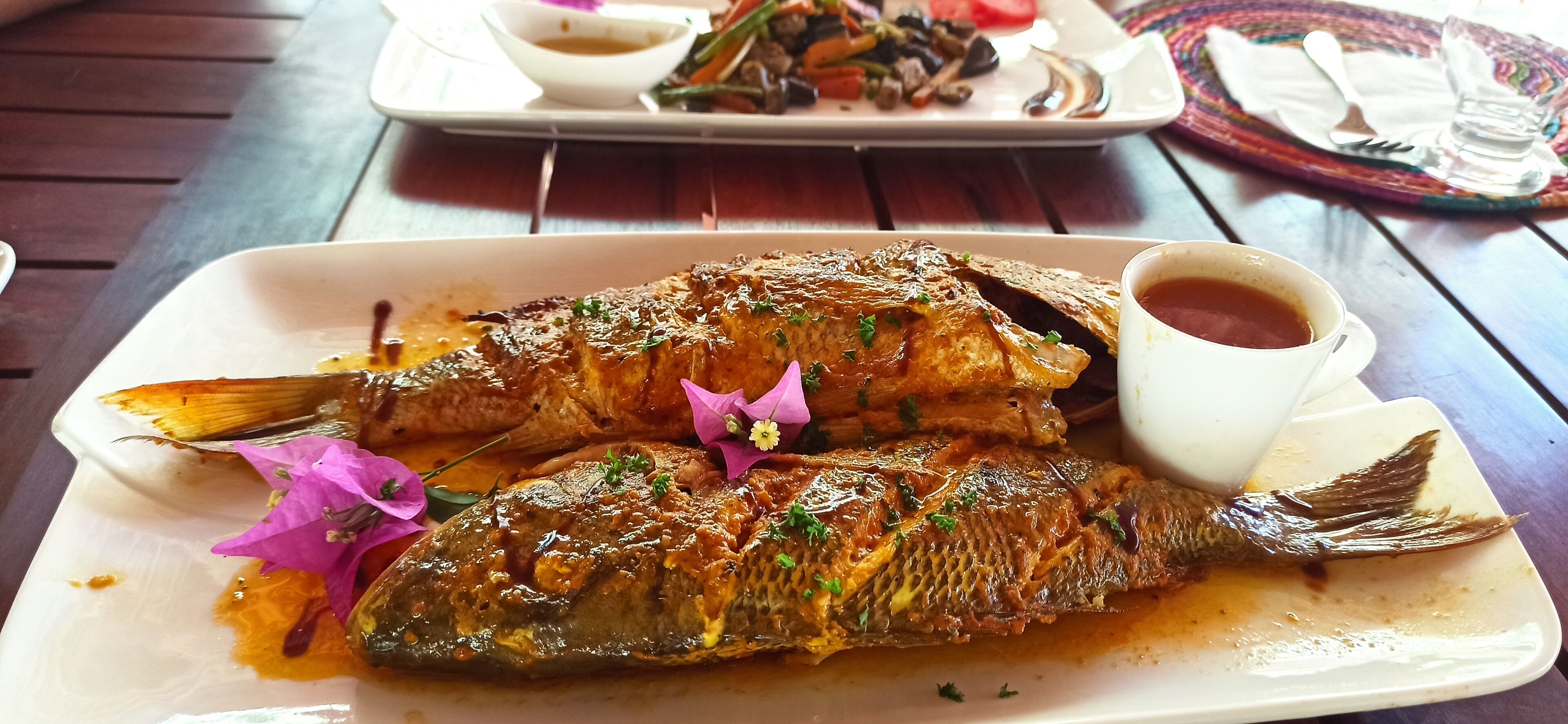 A plate of grilled fish with sauce and garnish, a cup of tea, and a side dish on a wooden table.