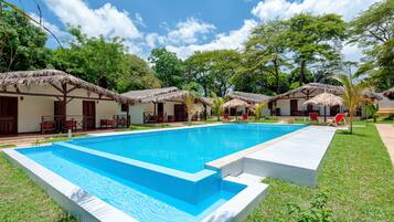 Una piscina rodeada de chozas con techo de paja, con mobiliario exterior y un exuberante paisaje verde.