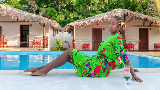A person relaxing by a pool with thatched-roof huts in the background. - BLUE SKY RESORT (Nosy Be)