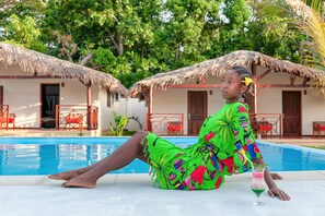 A person relaxing by a pool with thatched-roof huts in the background. - BLUE SKY RESORT (Nosy Be)