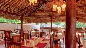 An outdoor dining area with wooden tables and chairs, thatched roof, and hanging light fixtures. - BLUE SKY RESORT (Nosy Be)
