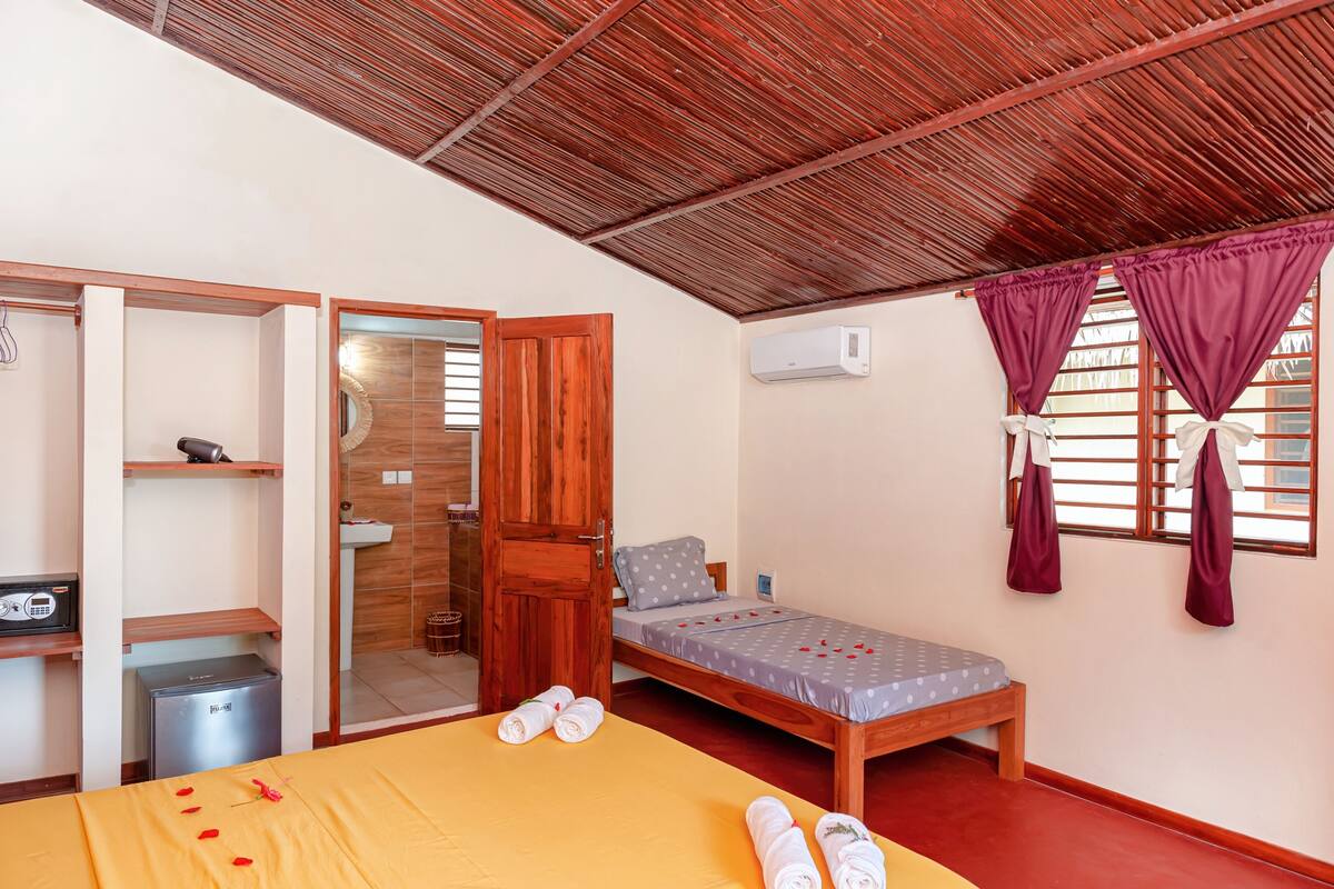 Um quarto individual com cama, escrivaninha pequena, espelho, guarda-roupa e banheiro visível por uma porta aberta.