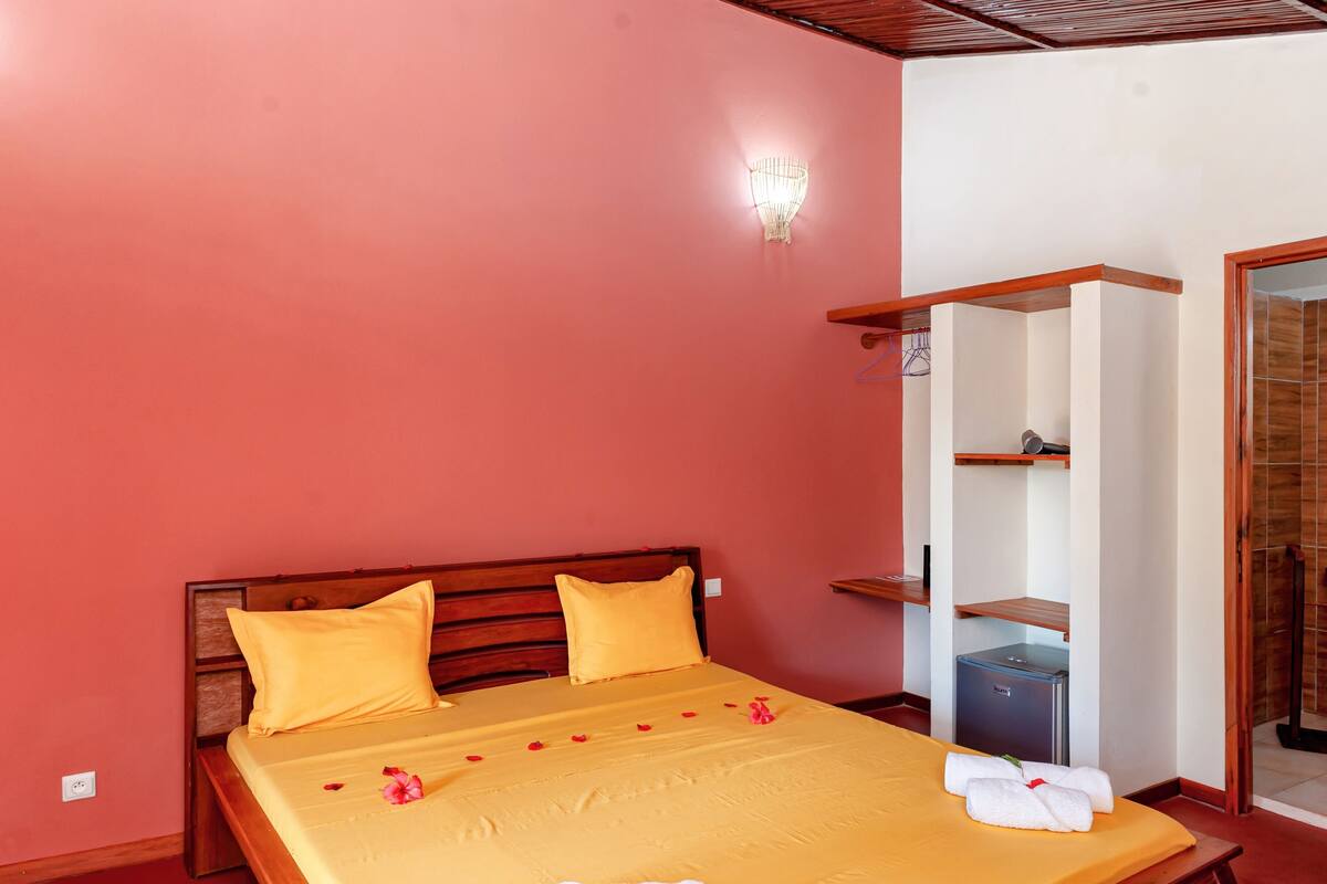 Quarto de hotel com cama, mesa de cabeceira, luminária na parede e um pequeno frigorífico.