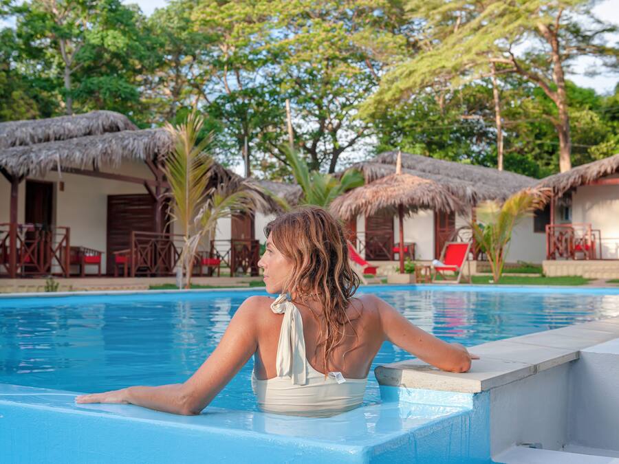A poolside view with a person relaxing by the pool edge, surrounded by thatched-roof huts and palm trees.