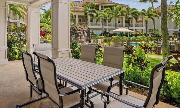 An outdoor patio with a table and chairs, surrounded by lush greenery and a pool.