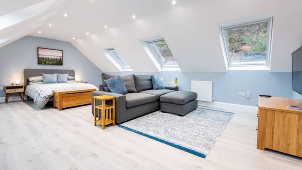 Une chambre spacieuse située sous les combles, avec un grand lit, un canapé d’angle gris, une table de chevet en bois et une petite table en bois. Elle est dotée de fenêtres de toit et d’une grande fenêtre qui laissent entrer la lumière naturelle.