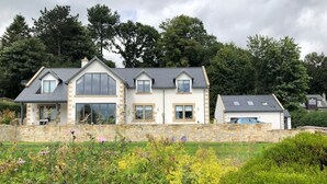 Une grande maison à deux étages, dotée de fondations en pierre, de nombreuses fenêtres et d’un jardin bien entretenu à l’avant.