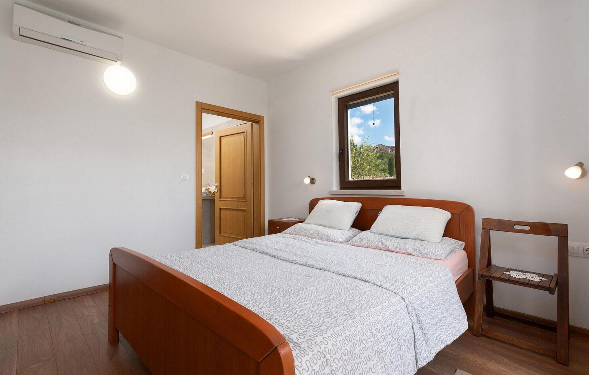 Un dormitorio con cama de madera, una mesita de noche, una pequeña mesa, una ventana con vistas y un equipo de aire acondicionado.
