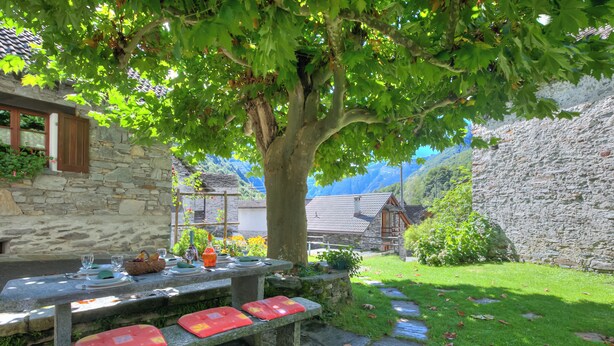 A stone table set outdoors with benches, surrounded by greenery and stone walls, overlooking a mountainous landscape. - Rustico al platano 2 (Sonogno)