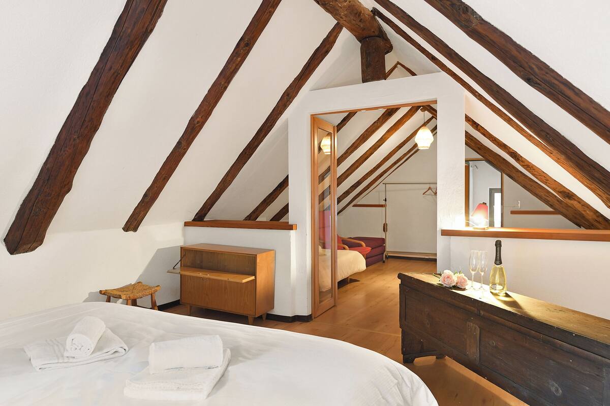 A bedroom with a sloped ceiling, wooden beams, a bed with white linens, a wooden dresser, and a mirror.