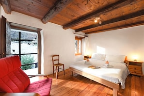 A bedroom with a wooden ceiling, a bed with white bedding, a red chair, a wooden nightstand, and a window with a view of greenery. - Rustico al platano 2 (Sonogno)