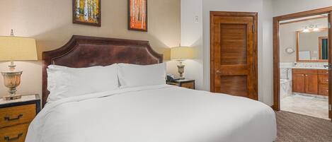 Habitación de hotel con una cama grande, dos lámparas de noche, un cabecero de madera, una mesita de noche, un aparador y un baño visible a través de una puerta abierta.