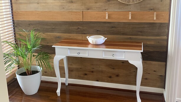 Une pièce avec un mur en bois, une table blanche avec des tiroirs, une plante en pot, un tableau représentant une plage et un miroir décoratif.