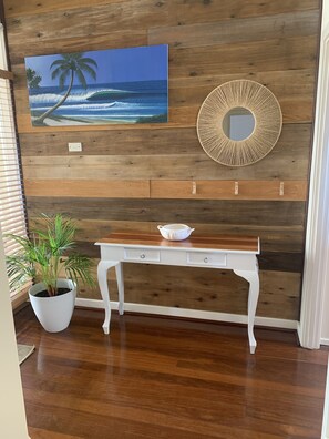 A room with a wooden wall, a white table with drawers, a potted plant, a painting of a beach, and a decorative mirror. - Great central location, one street back from the beach. (Beresford)