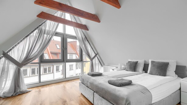 A bedroom with a large window, a bed with grey bedding, and wooden beams on the ceiling. - MARGI Gran Apartament (Szczecin)
