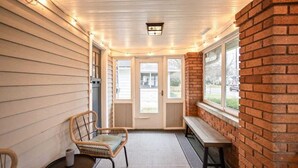 A covered porch with wicker chairs, a bench, and a brick wall. - Backyard+Porch, Pet-Friendly | Broad Ripple Stay (Indianapolis)