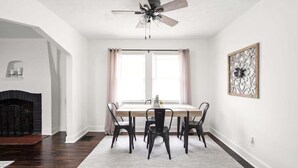 A dining area with a table, chairs, a ceiling fan, a fireplace, and a decorative wall piece. - Backyard+Porch, Pet-Friendly | Broad Ripple Stay (Indianapolis)