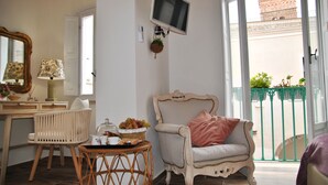 Une pièce avec un fauteuil en osier, une table sur laquelle se trouve un panier de fruits, un téléviseur fixé au mur et une vue sur un balcon agrémenté de plantes.