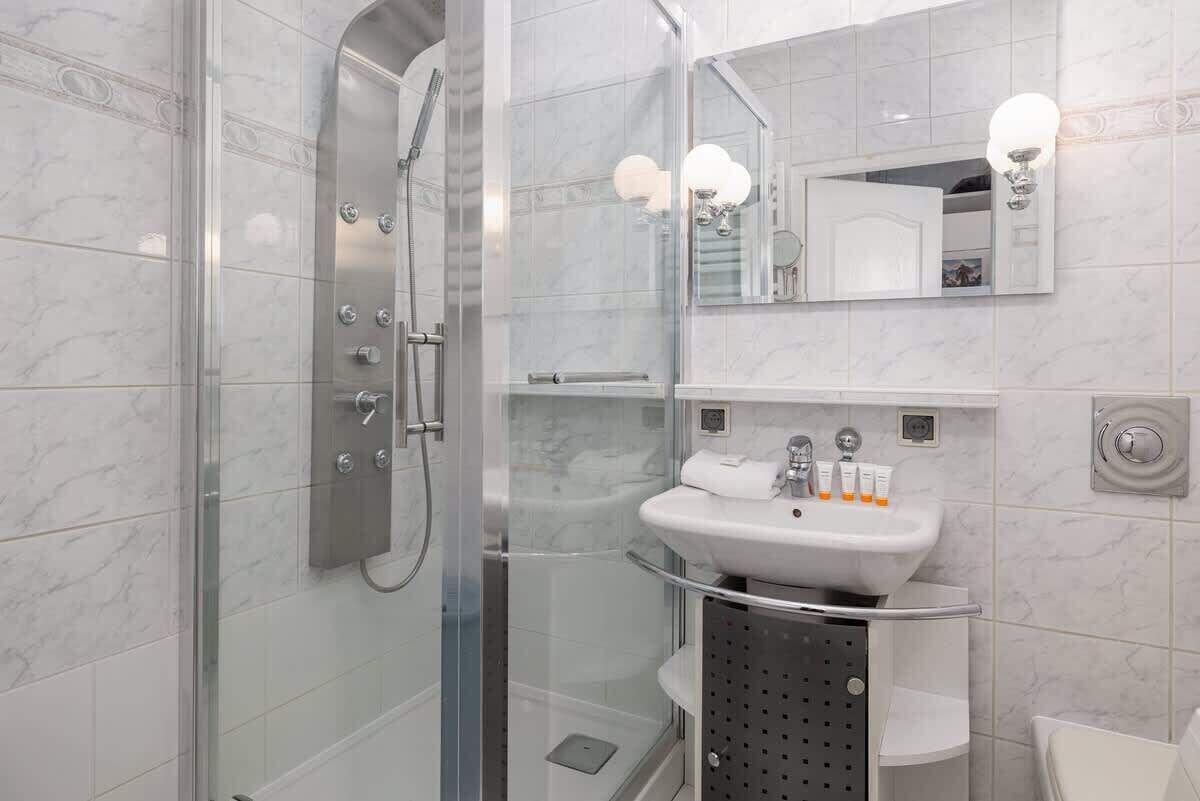 Een moderne badkamer met een douchecabine van glas, een wit wastafelmeubel met een glazen plank en een grote spiegel met wandlampen.