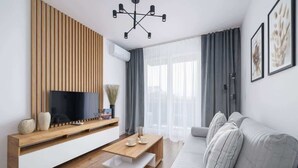 A modern living room with a wooden TV stand, a flat-screen TV, a wooden coffee table, a grey sofa, and a large window with sheer curtains.