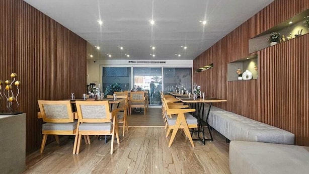 A modern restaurant interior with wooden panel walls, a central dining area with wooden tables and chairs, and a seating area with cushioned benches. - Nest By Rivido Apart Hotel (Bengaluru)