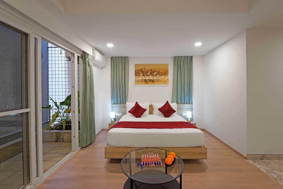 A modern hotel room with a large bed, a glass coffee table with chess pieces and oranges, and a view of a balcony with plants.