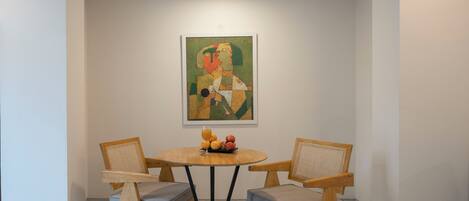 A dining area with a round table, two wooden chairs with cushions, a painting on the wall, and a glass of orange juice on the table.