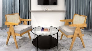 A modern living room with a flat-screen TV mounted on a white wall, two wooden chairs with rattan backs, and a glass coffee table with a chess set. - Nest By Rivido Apart Hotel (Bengaluru)